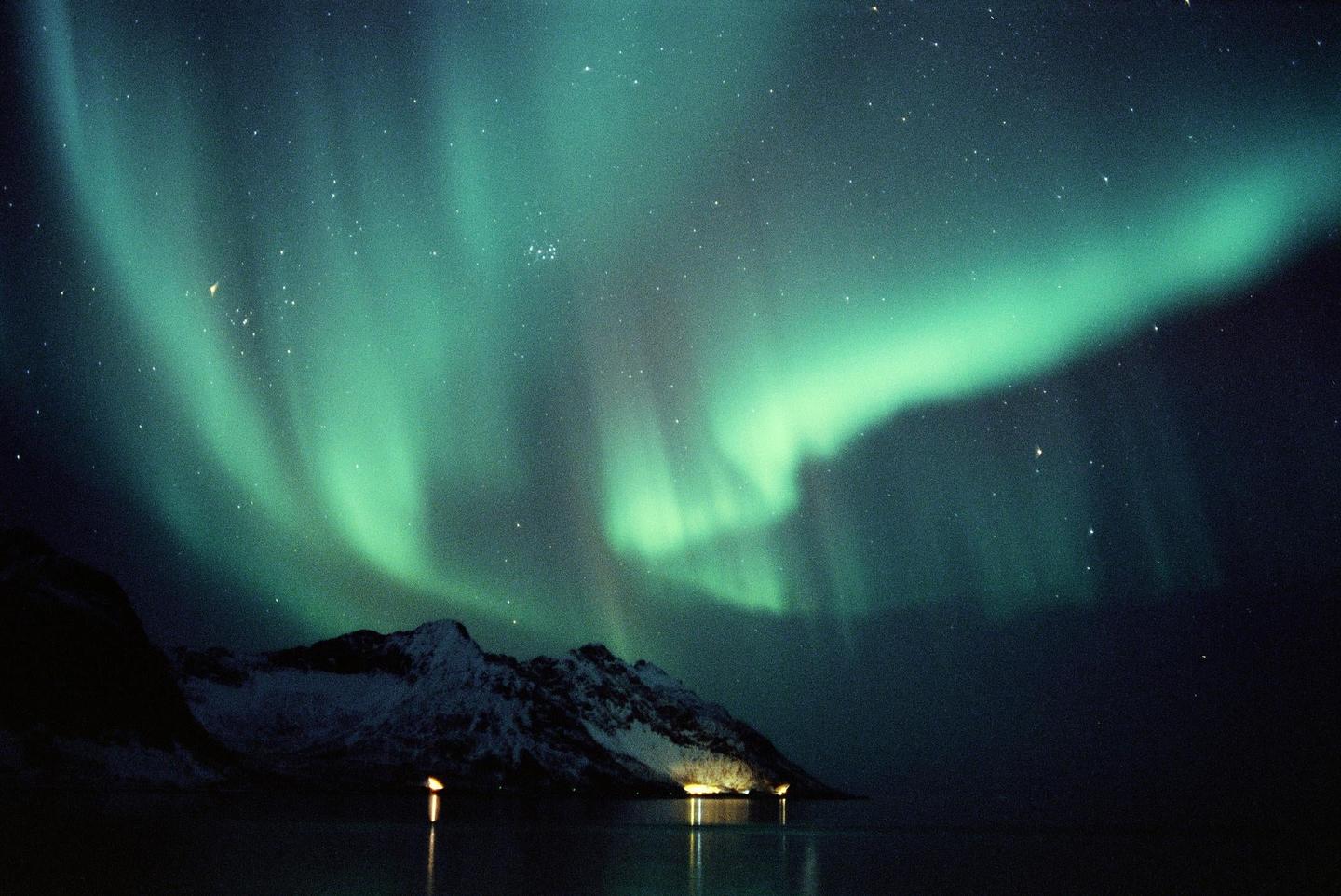 The aurora borealis glows green above a snowy mountain and a body of water with lights reflecting on its surface.