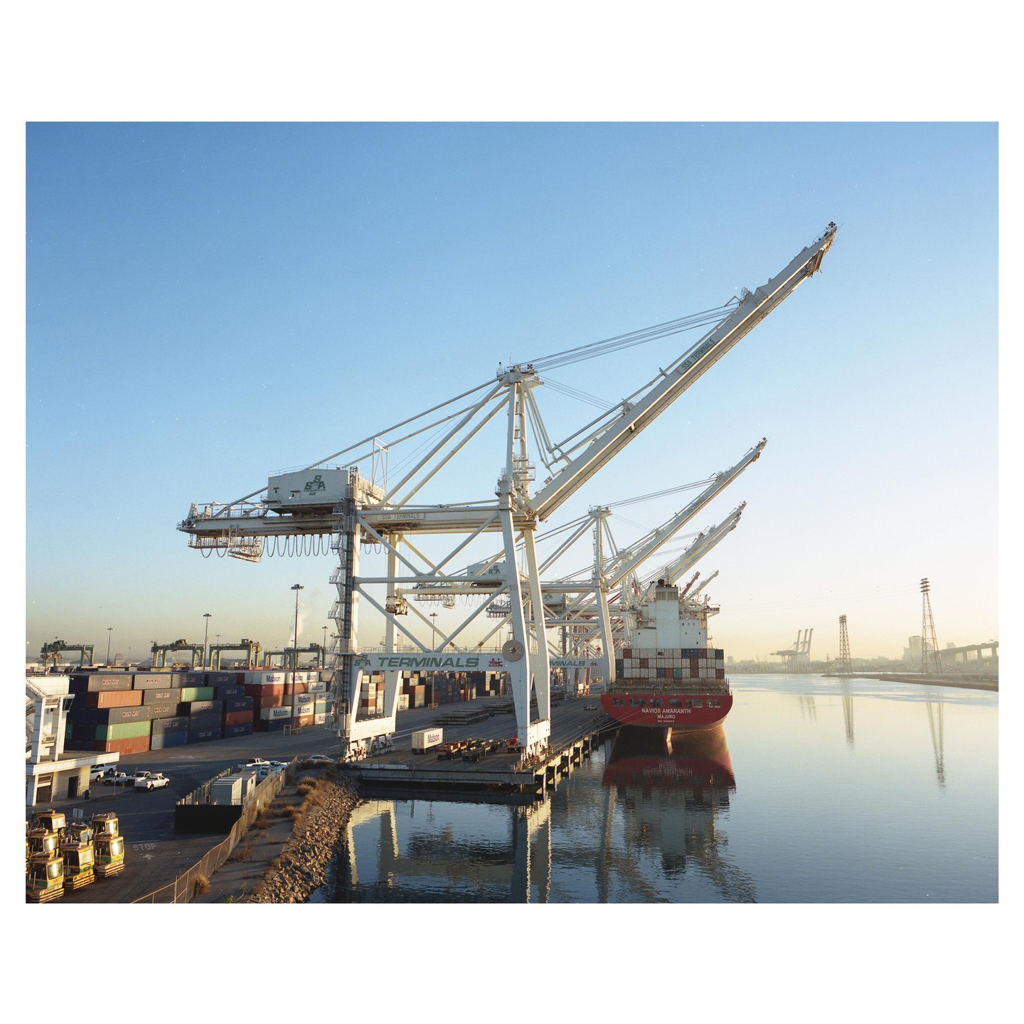 A large red cargo ship is docked at a terminal with several cranes under a clear sky.