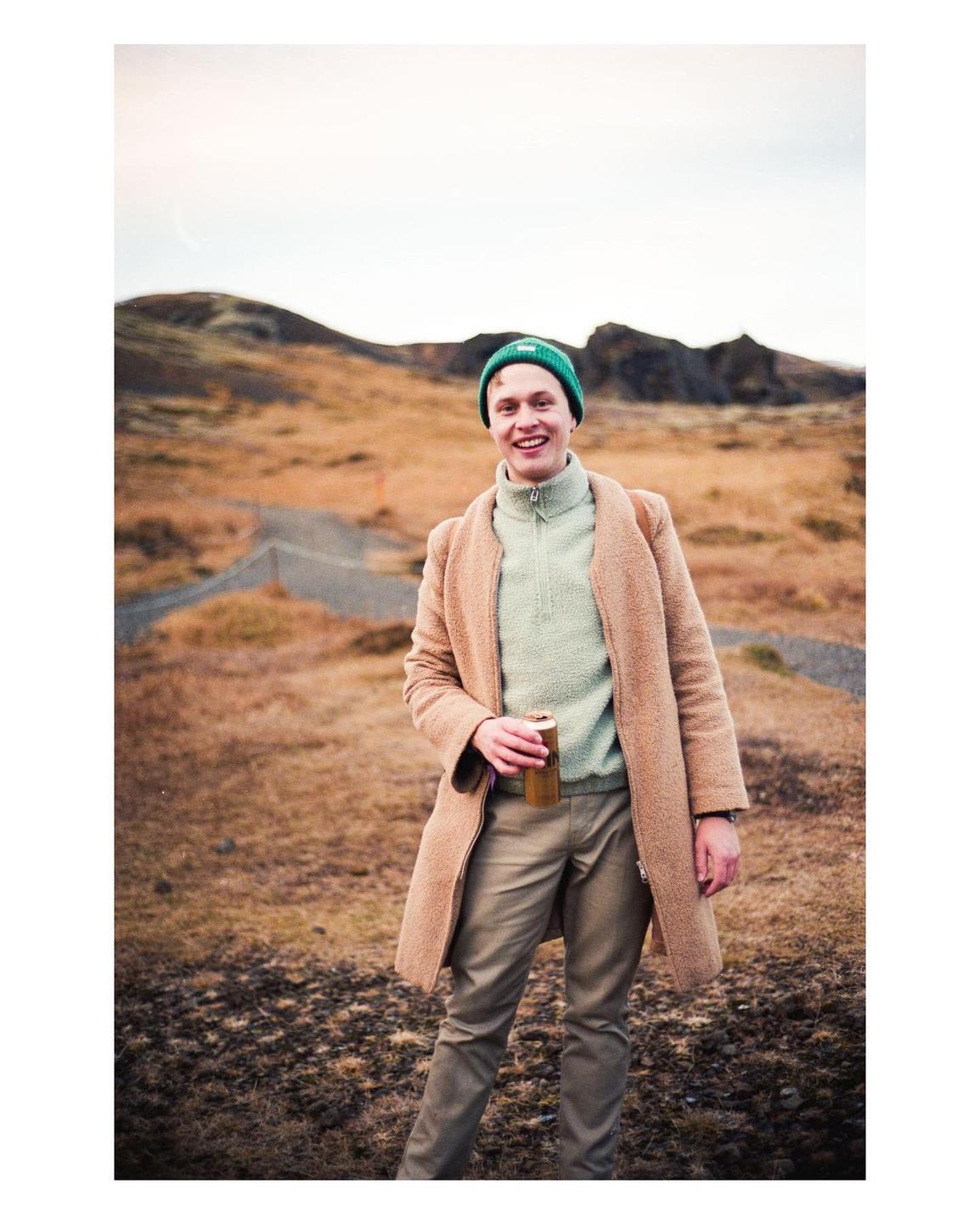 A man in a green beanie and a brown coat smiles while holding a can in a barren, hilly landscape.
