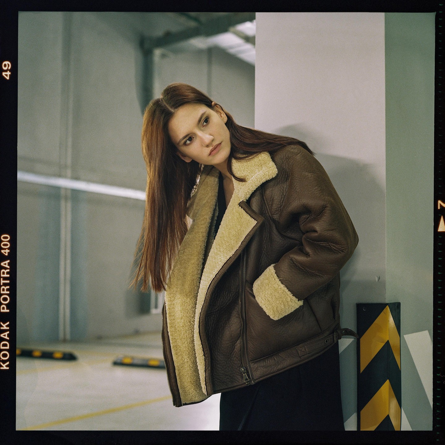 A woman in a brown and cream jacket leans against a pillar in a parking garage.