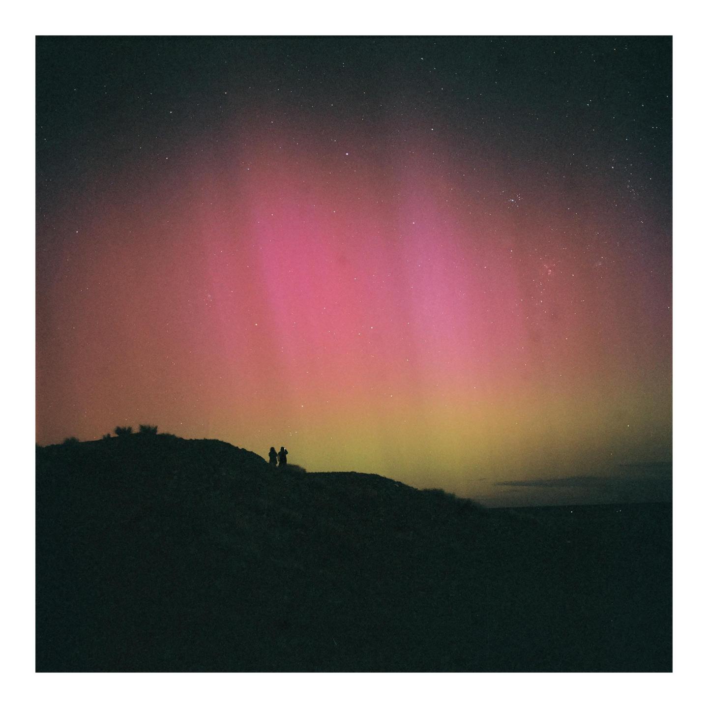 Two people stand on a dark hill silhouetted against the vibrant pink and yellow hues of the aurora australis and a starry night sky.