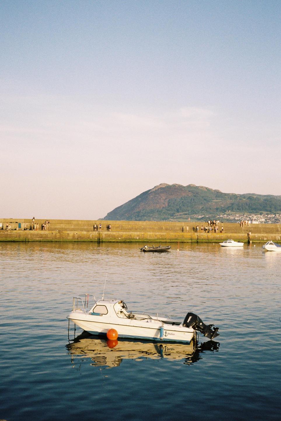 A small white boat with an orange buoy floats on the water in a marina with a concrete pier and a mountain in the background.