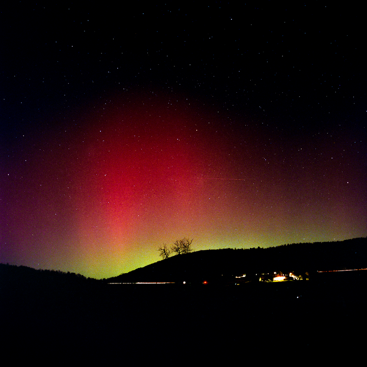 The northern lights glow red and green over a dark landscape with a few distant lights and a starry sky.