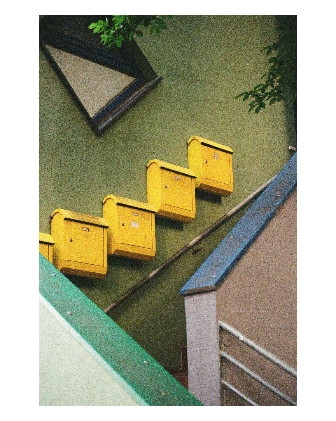 Four yellow mailboxes are mounted on a green wall next to a building with a blue roof.