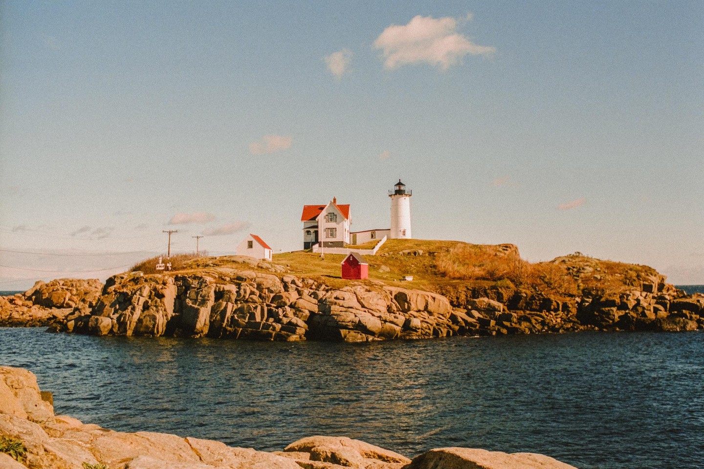 A white lighthouse and house with a red roof stand on a rocky island surrounded by water.