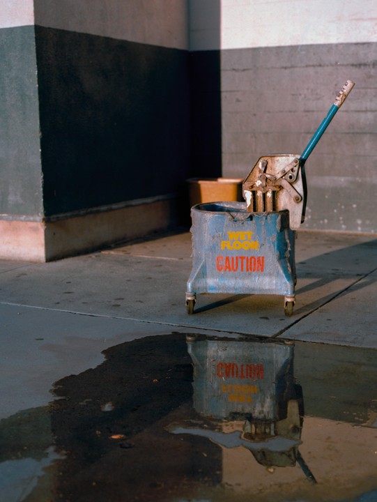 A mop bucket with "CAUTION WET FLOOR" written on it stands next to a puddle of water, reflecting the text.