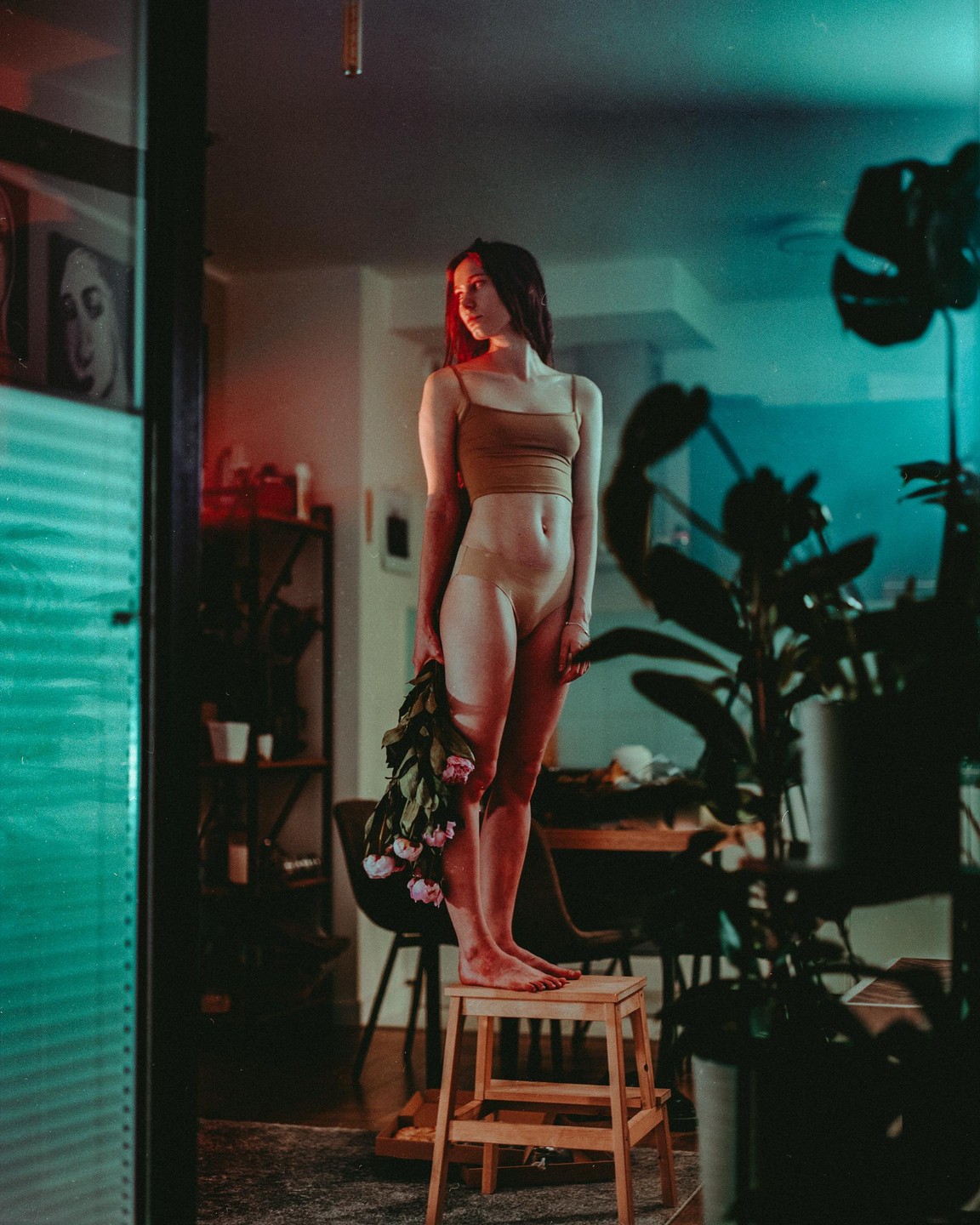 A woman stands barefoot on a stool holding a bouquet of flowers in a room with colorful lighting.