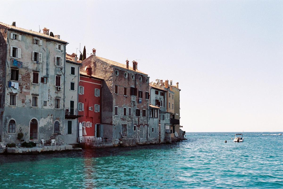 A row of colorful buildings with shuttered windows stands along the edge of a bright blue sea with a small boat.