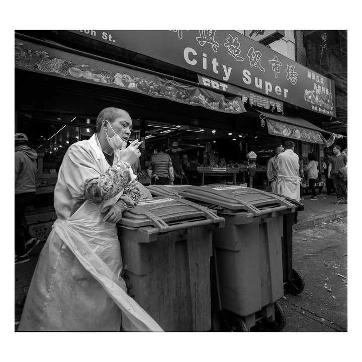 A man in an apron and face mask smokes a cigarette next to two trash cans in front of a City Super grocery store.