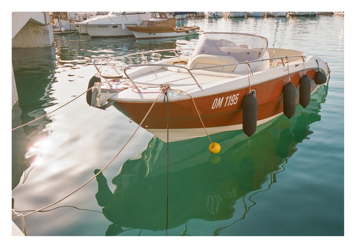 A small boat with a brown hull and white deck is moored in a harbor, with other boats visible in the background.