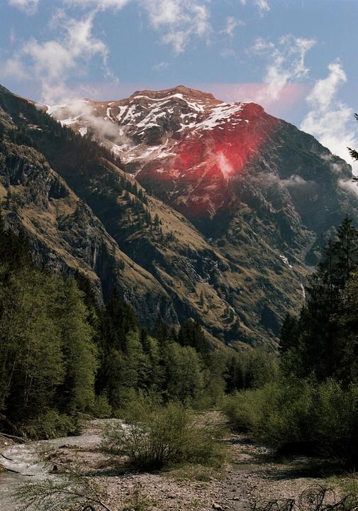 A mountain with snow on its peak and a red light leak is visible above a forest and a river.