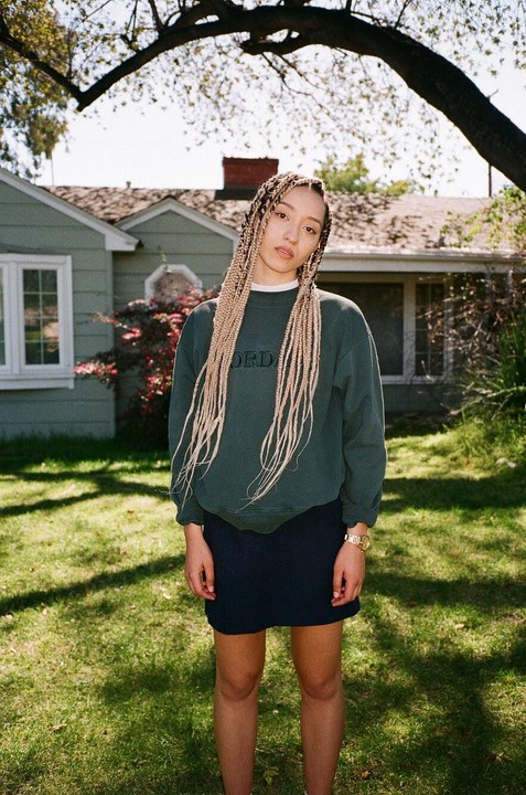 A woman with long braids stands in a grassy yard in front of a house.