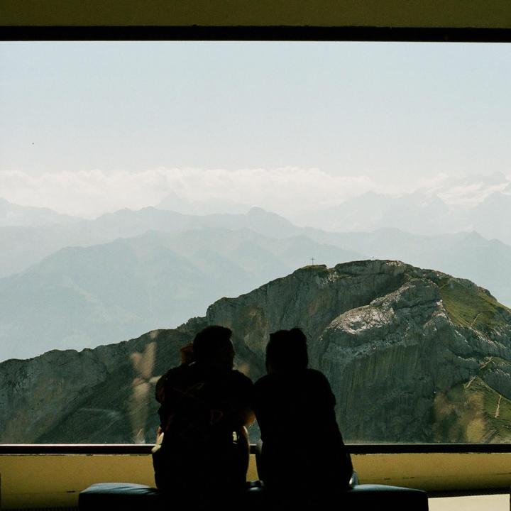 Two people sit with their backs to the viewer, looking out a window at a mountain range with a cross on the peak.