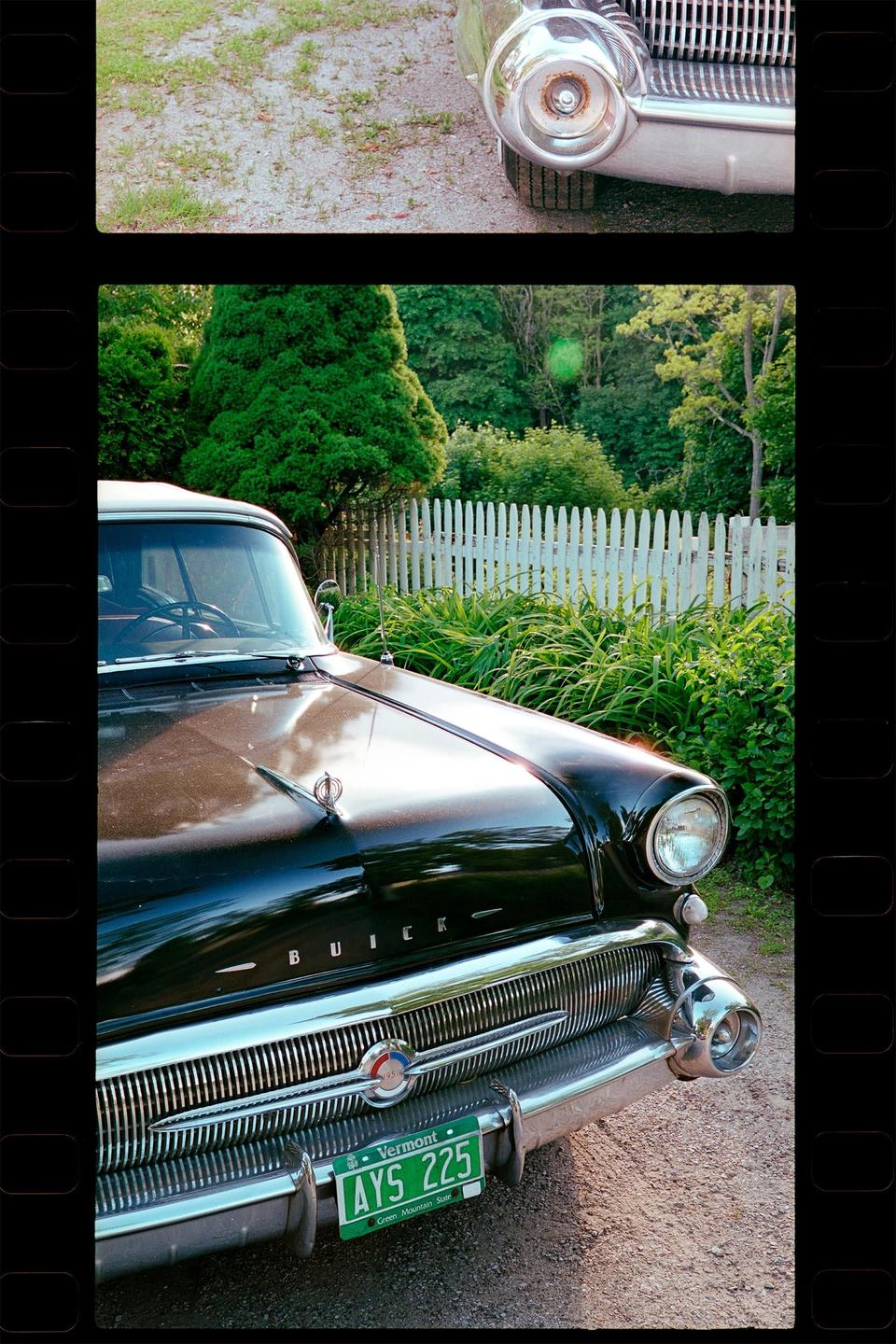 A black 1957 Buick is parked on a gravel driveway next to a white picket fence and lush green foliage.