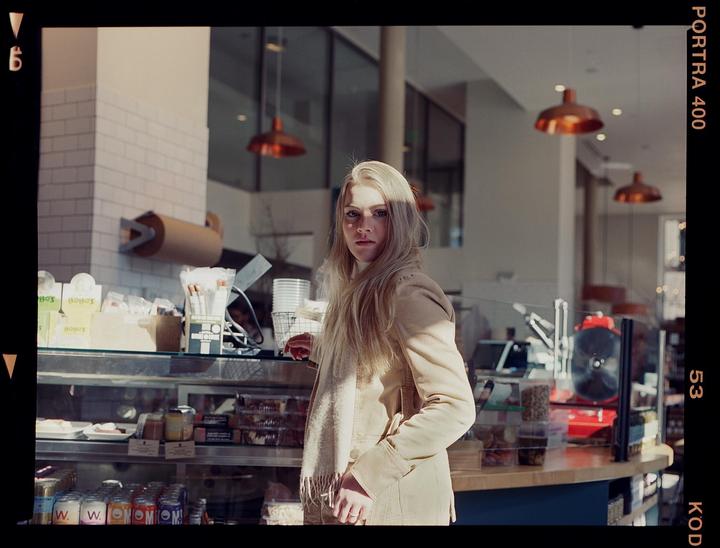 A woman stands in a cafe with a display case of food and drinks in front of her.