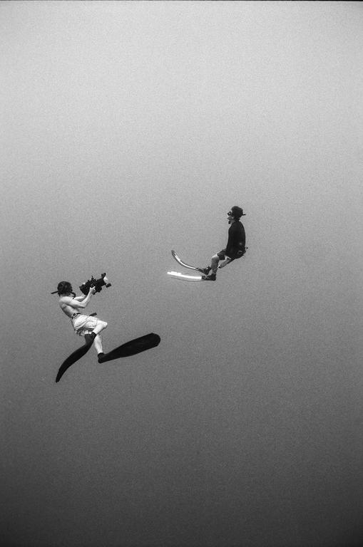Two men are underwater, one holding a camera and the other on a hydrofoil.