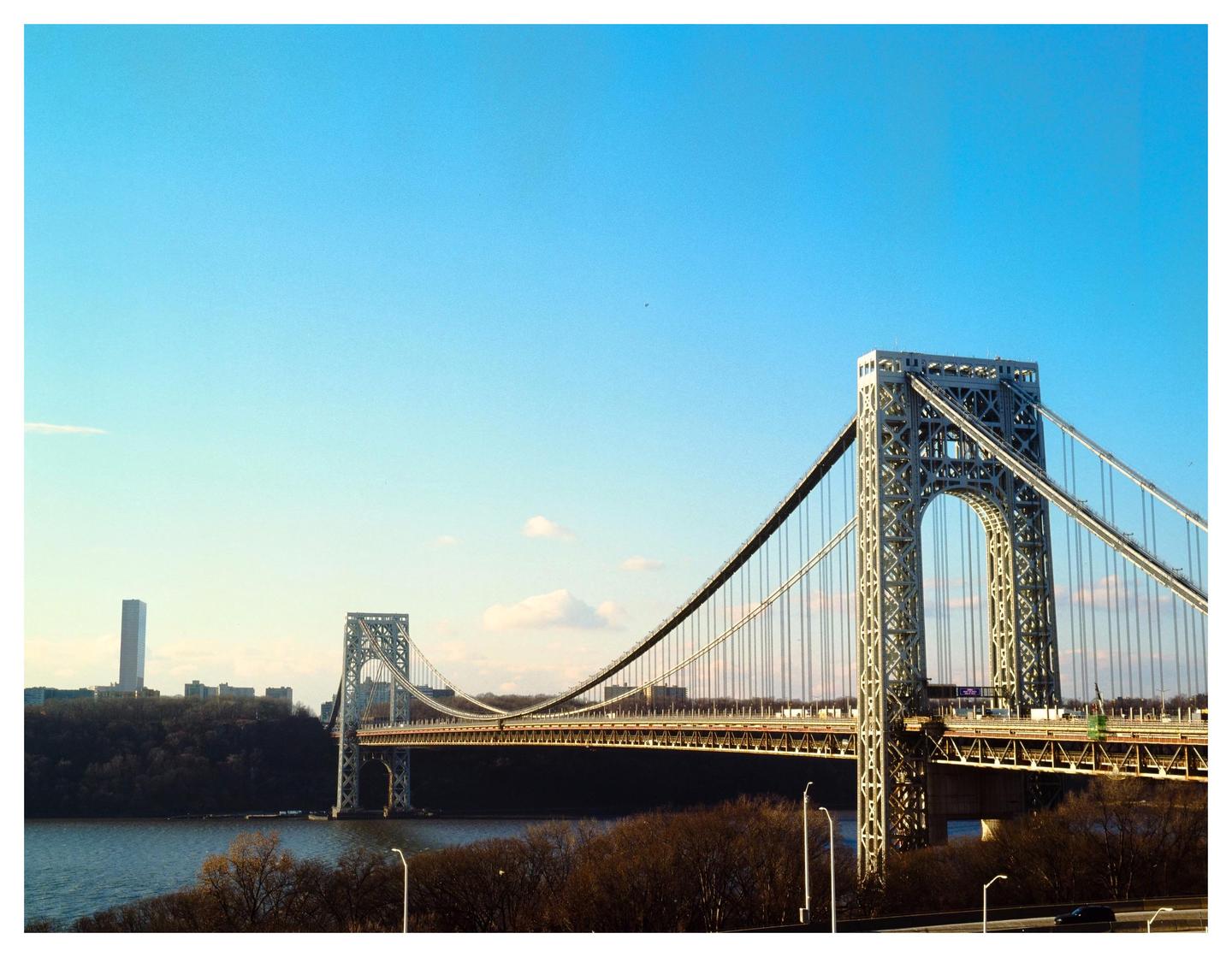 A suspension bridge spans a river with a city skyline and trees on the far bank under a clear sky.