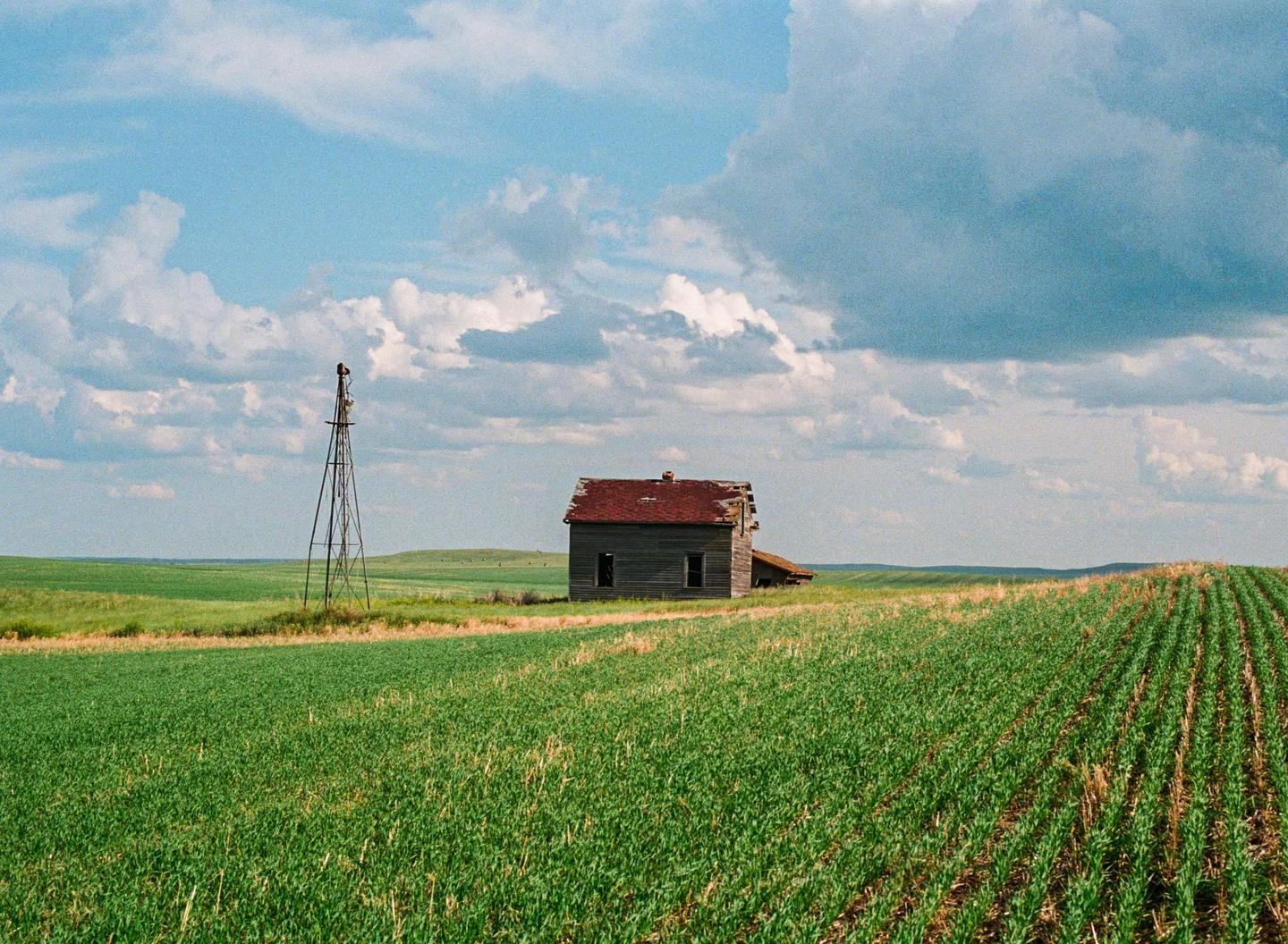 An abandoned house with a red roof stands next to a windmill in a vast green field under a cloudy sky.