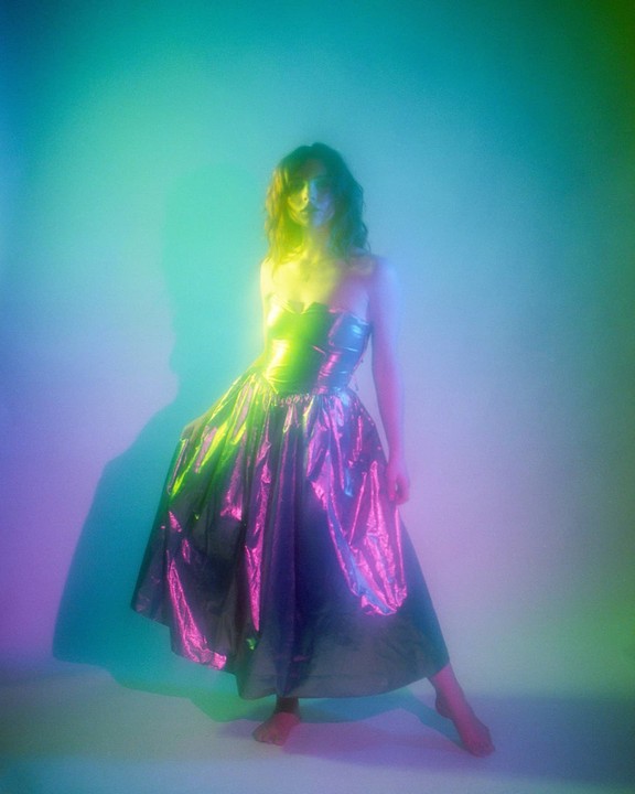 A woman in a shiny dress stands barefoot in a room lit with colorful neon light.