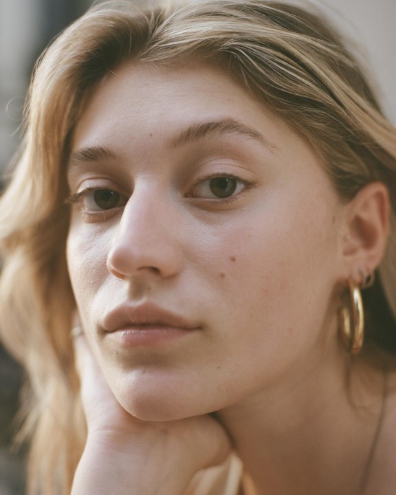 A woman with blonde hair and gold hoop earrings rests her chin on her hand while looking directly at the viewer.