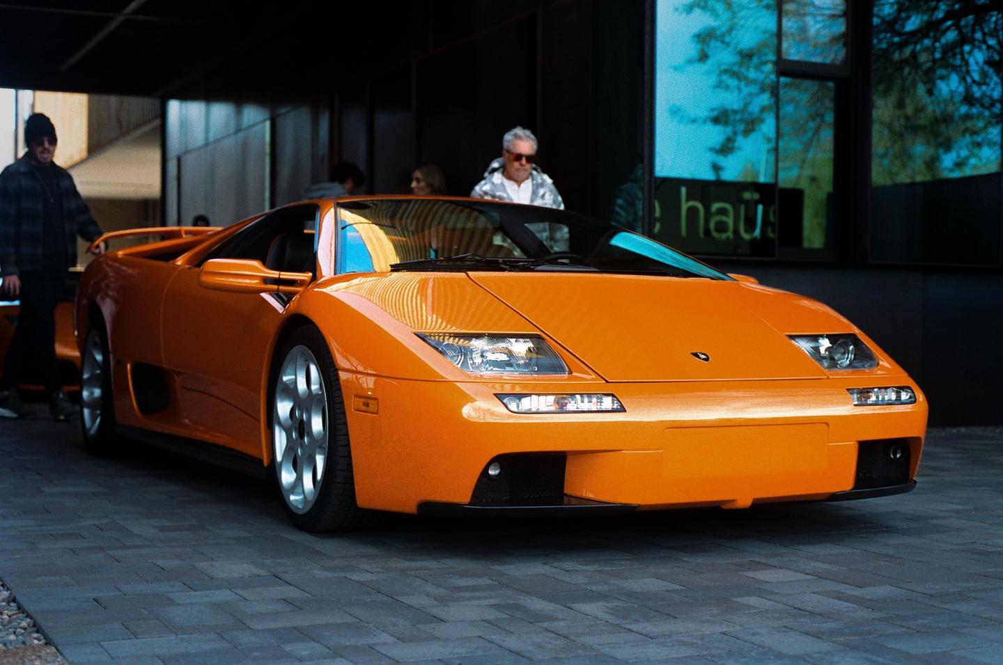 An orange sports car is parked on a paved surface with people standing in the background.