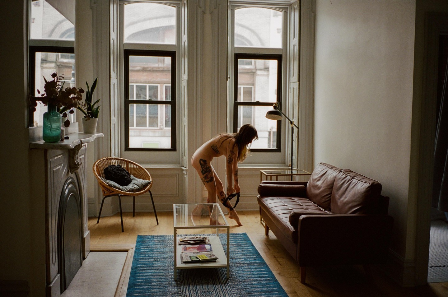 A woman bends over in a living room with a brown couch, a glass coffee table, and a blue rug.