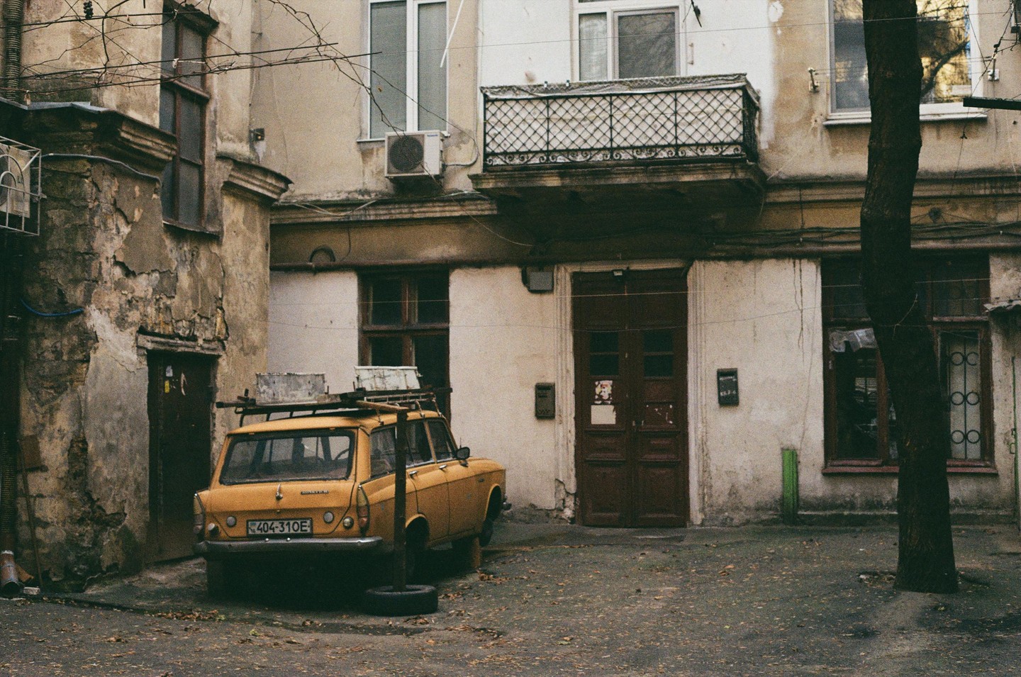 A yellow station wagon with a roof rack is parked in a narrow alleyway between old, weathered buildings with a tree trunk on the right.