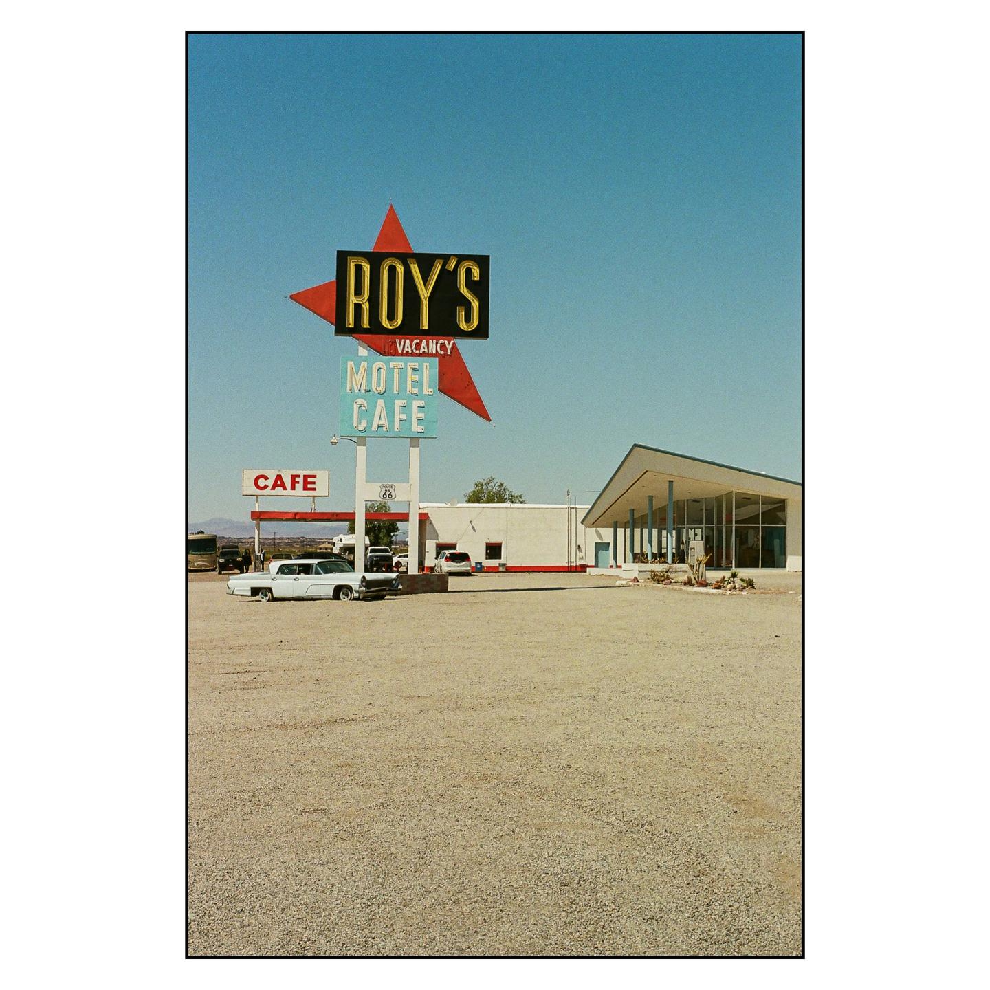 A large sign for Roy's Motel and Cafe stands in a gravel lot with a building and cars behind it.