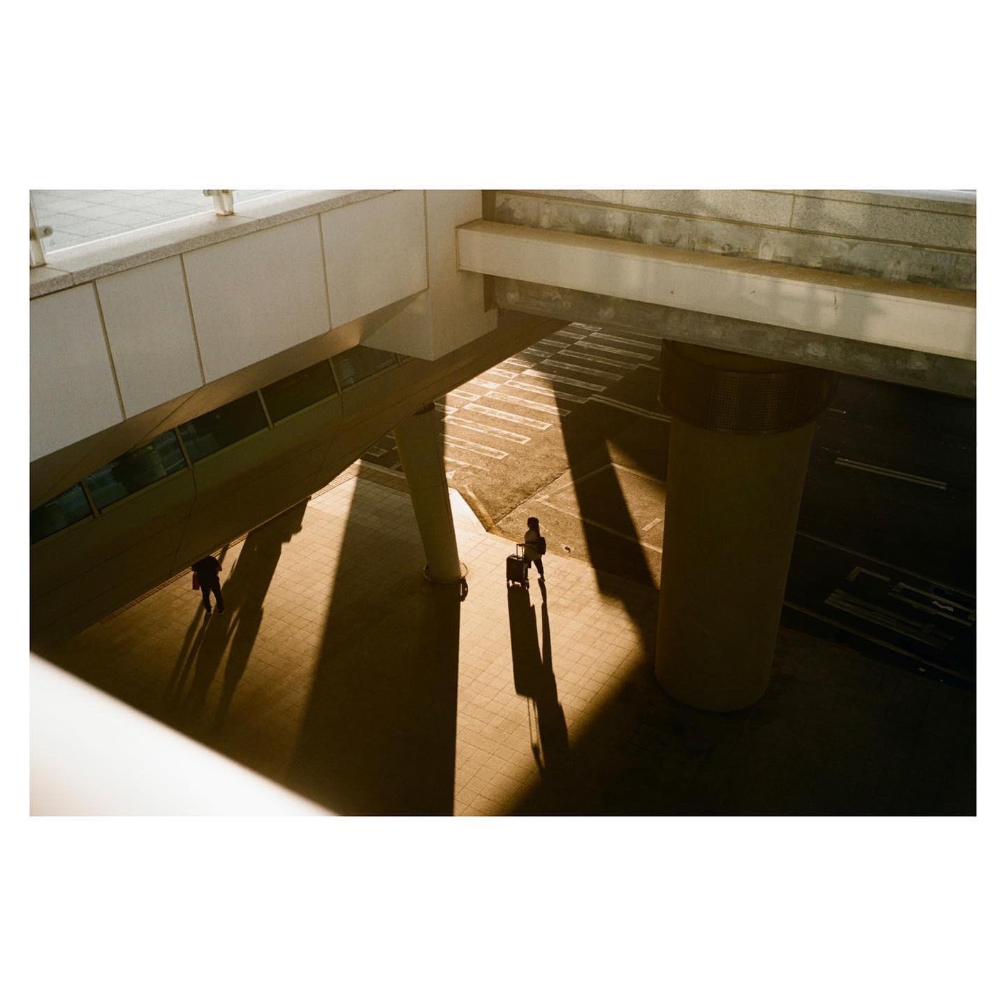 Two people walk on a sunlit street under an overpass, casting long shadows.