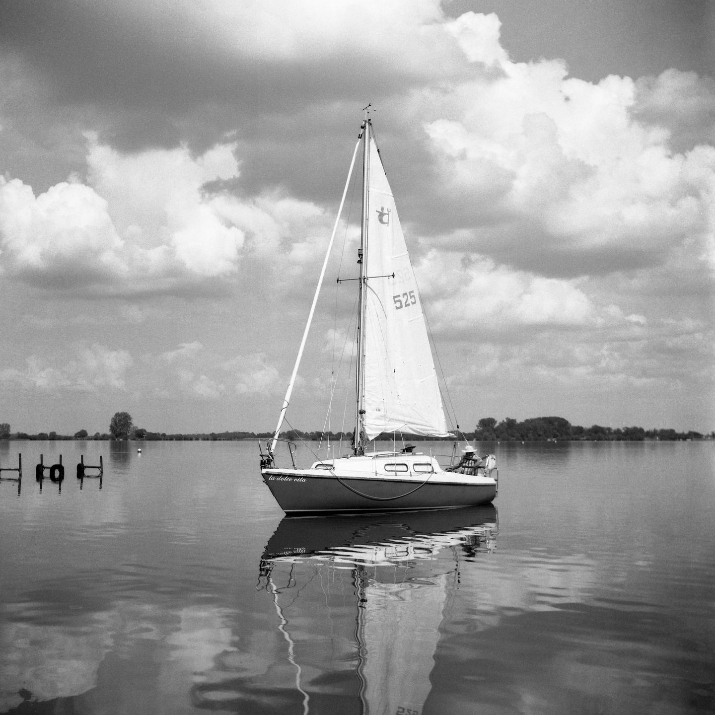 A sailboat with its sail up is reflected in the still water of a lake under a cloudy sky.