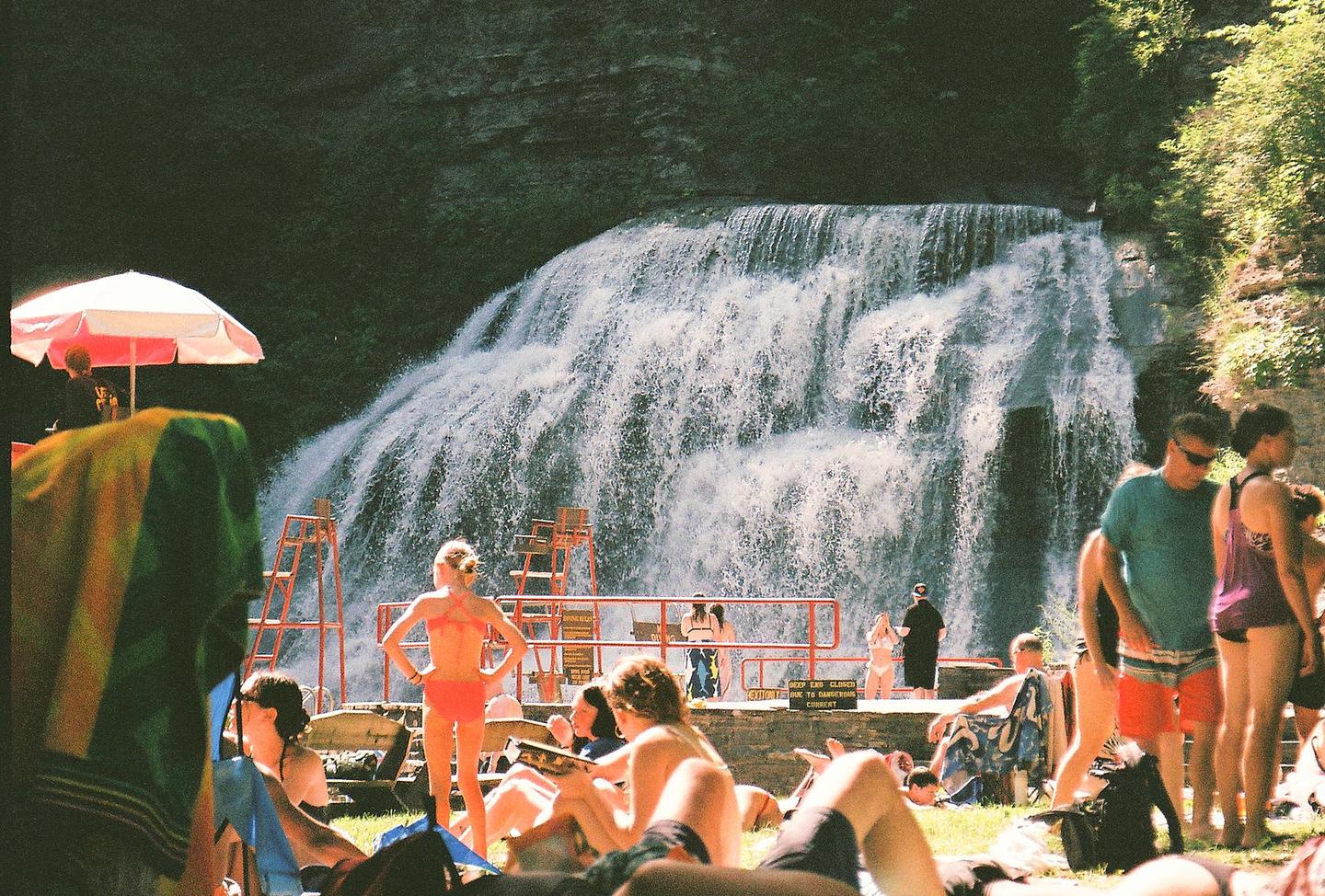 A crowd of people relax on the grass in front of a large waterfall with several lifeguard chairs and a red railing.