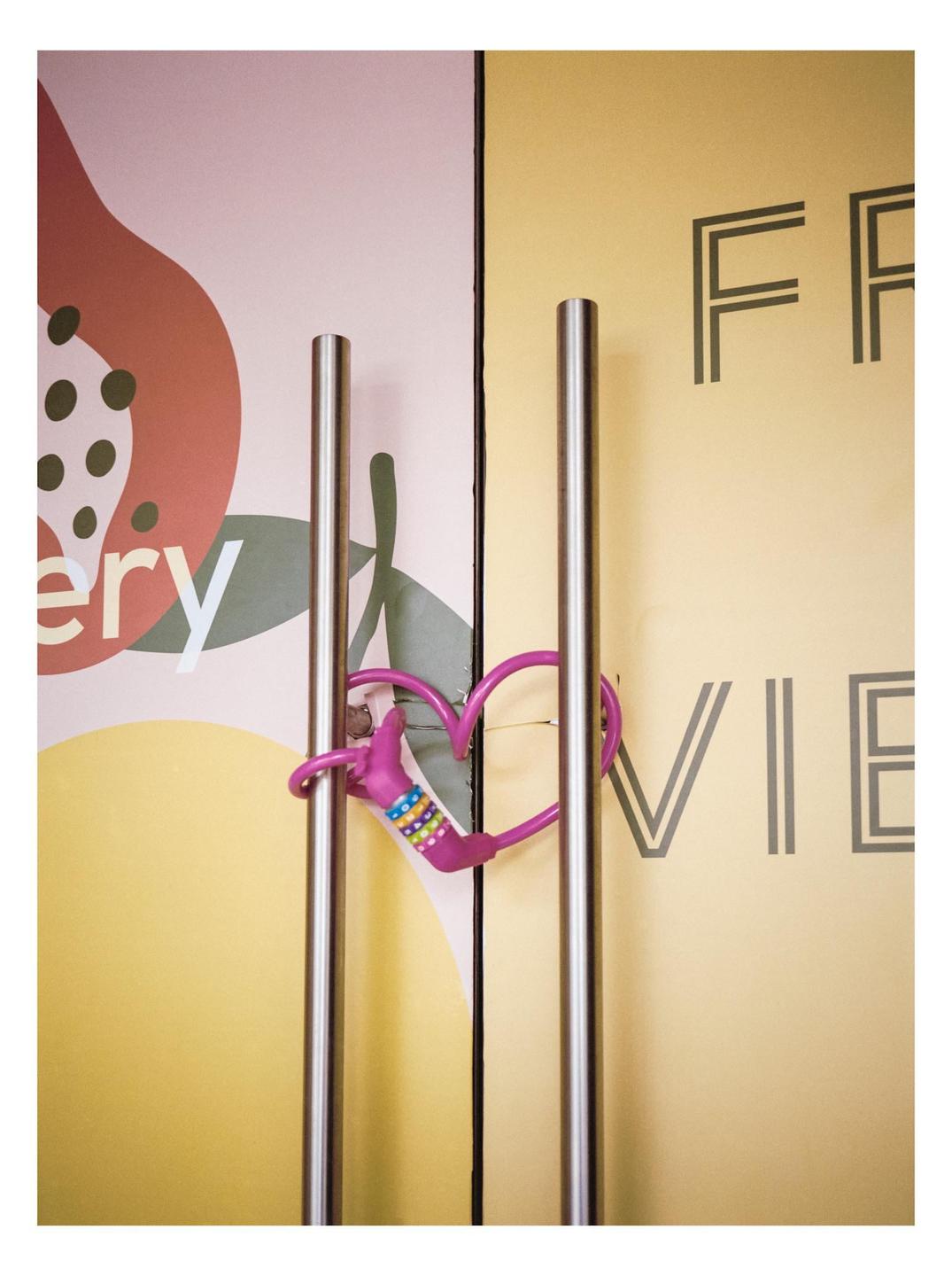 A pink heart-shaped lock is attached to two metal door handles in front of a colorful wall with text.