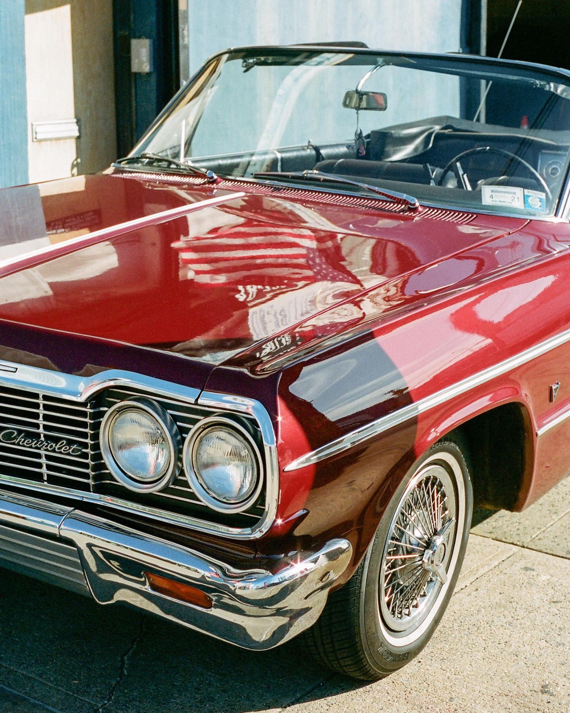 A red Chevrolet convertible is parked on a street with an American flag reflected on its hood.