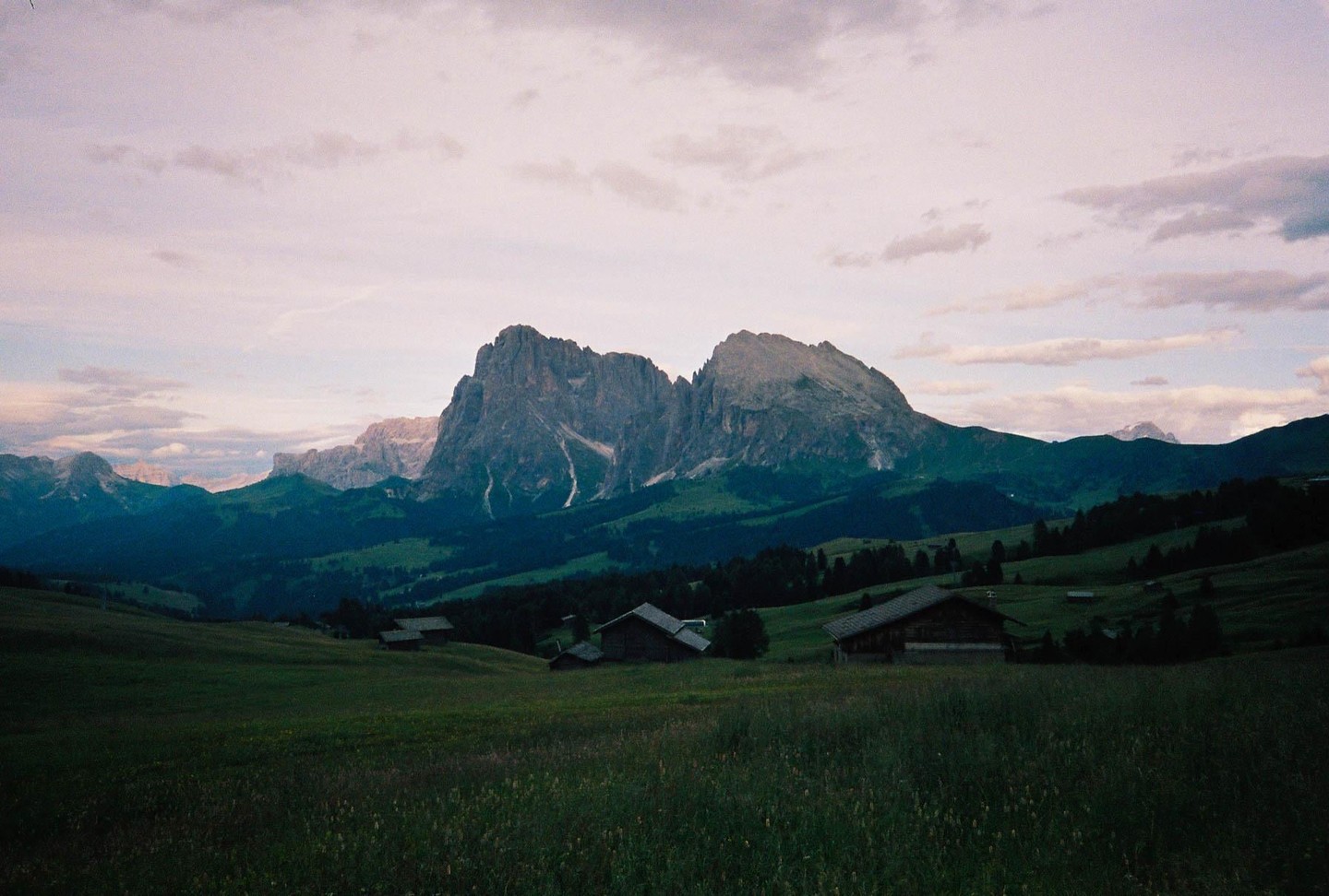 A mountain range rises above a valley with scattered wooden houses and green fields under a cloudy sky.