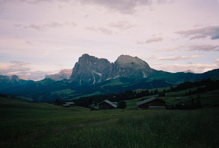 A mountain range rises above a valley with scattered wooden houses and green fields under a cloudy sky.