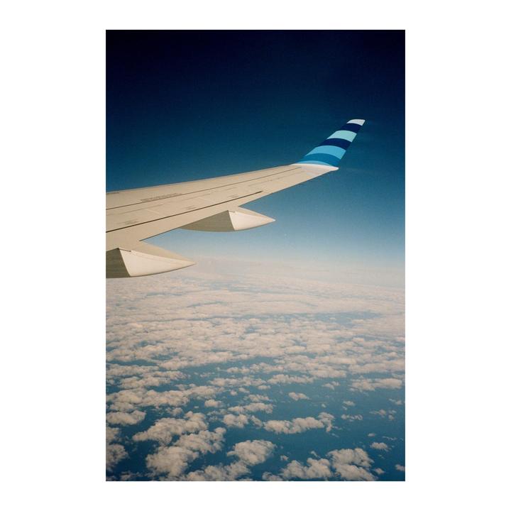 An airplane wing is visible above a layer of clouds with a clear blue sky above.