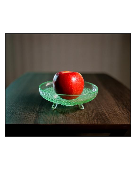 A red apple sits in a green glass bowl on a wooden table.