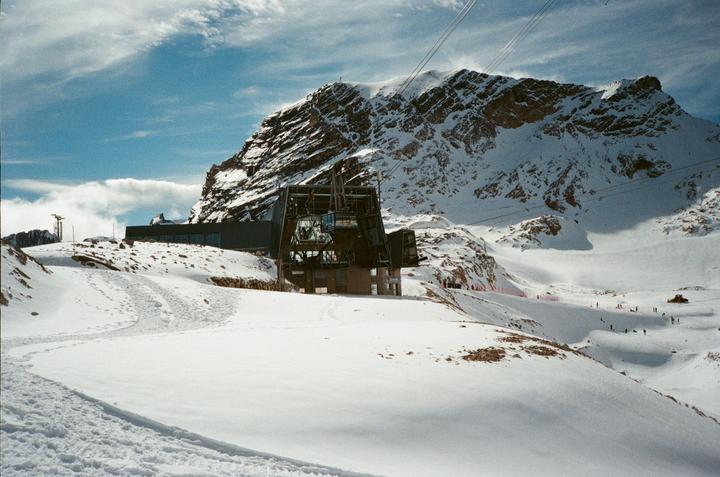 A ski resort with a gondola station is nestled in a snow-covered mountain landscape under a blue sky.