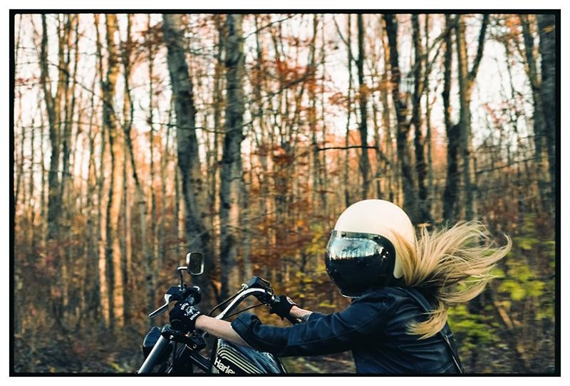 A woman with long hair and a helmet rides a motorcycle through a forest with autumn foliage.