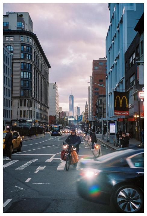 A man on a bicycle with bags on the handlebars rides down a city street with tall buildings and a McDonald's sign.