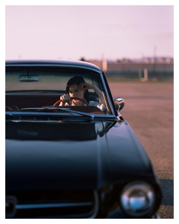 A woman sits in a black car with a red steering wheel, looking directly at the viewer.