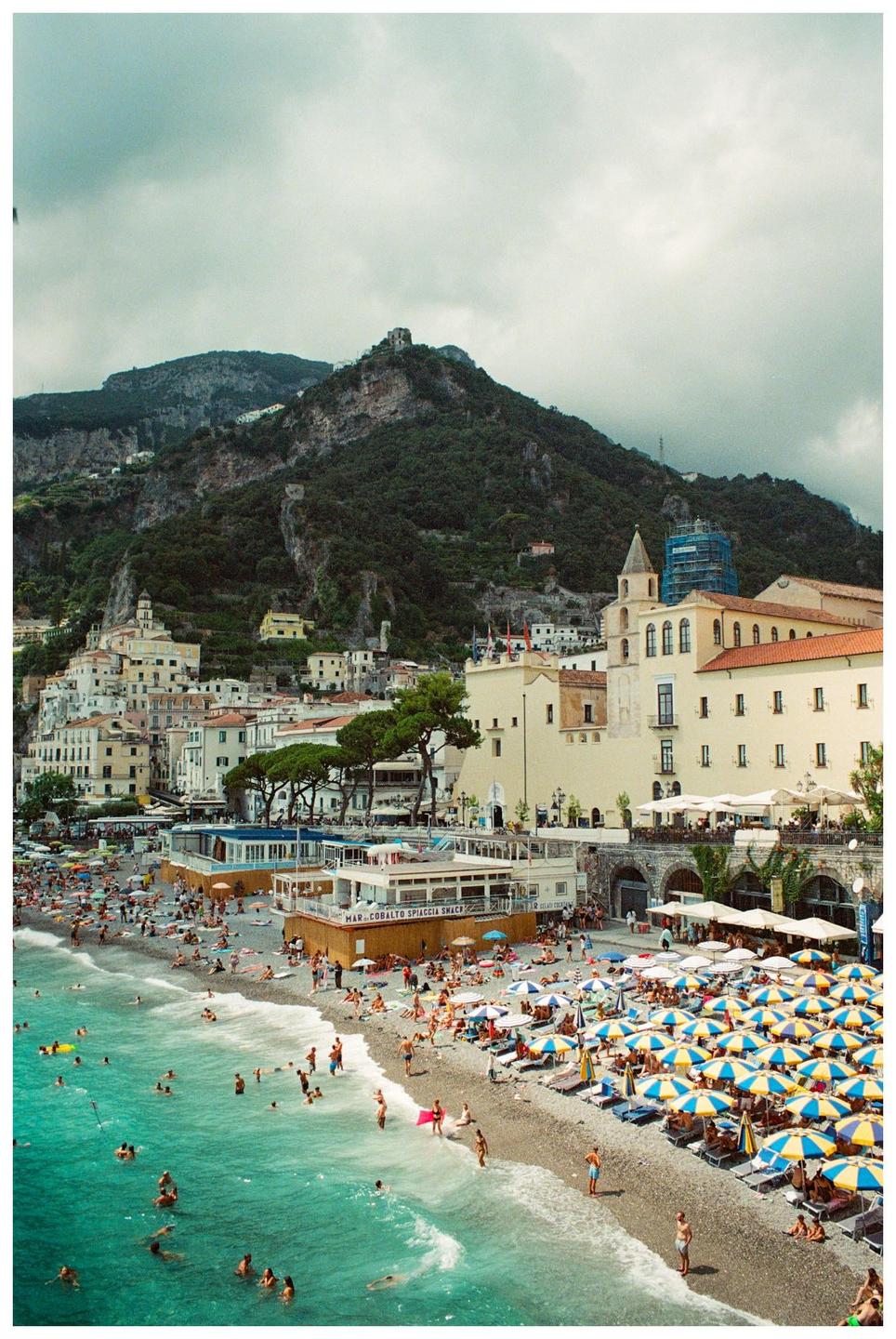 A crowded beach with many people swimming and sunbathing is nestled at the base of a mountain town with buildings climbing up the hillside.