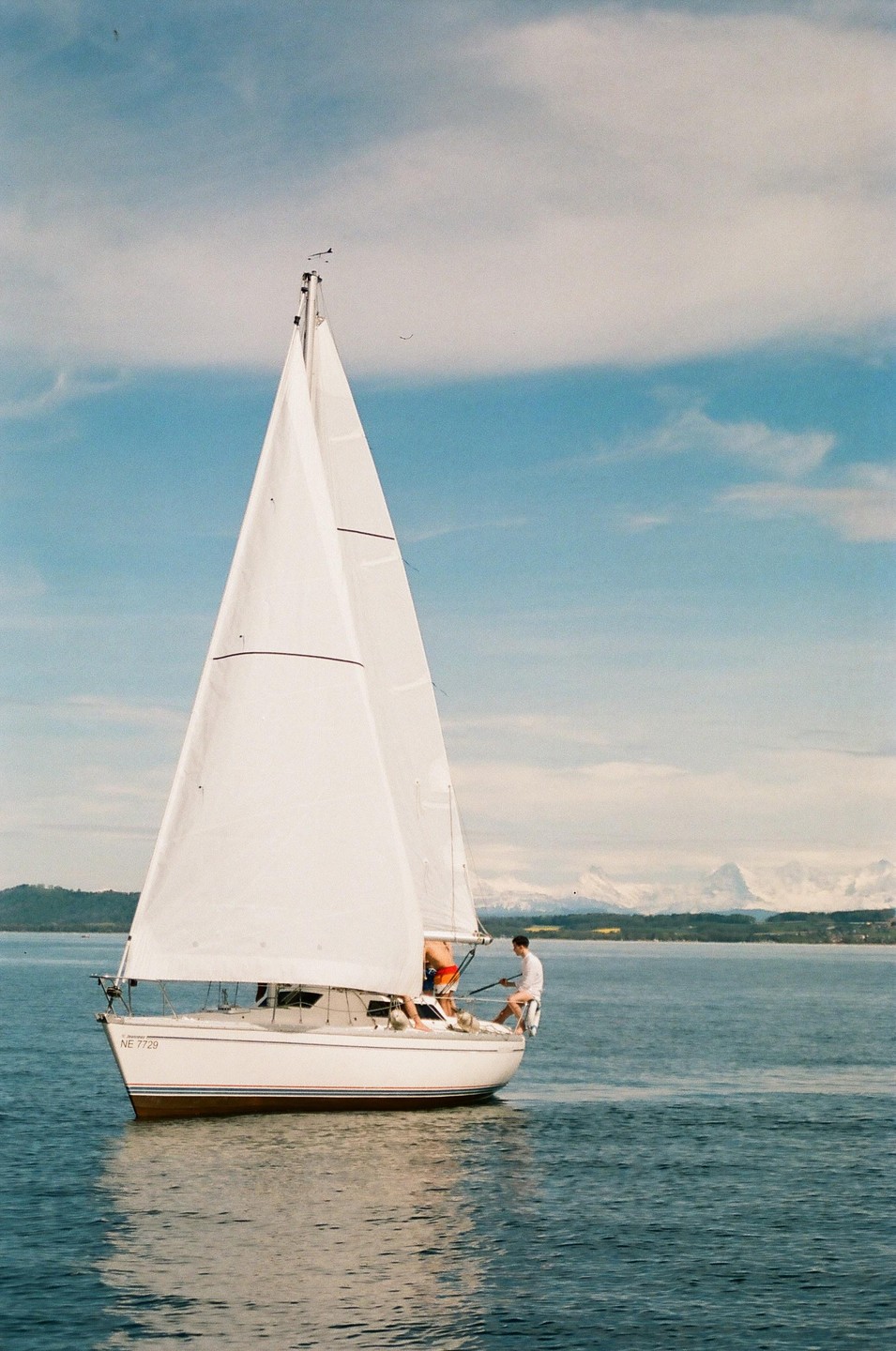 A sailboat with white sails glides across a body of water with mountains in the distance.