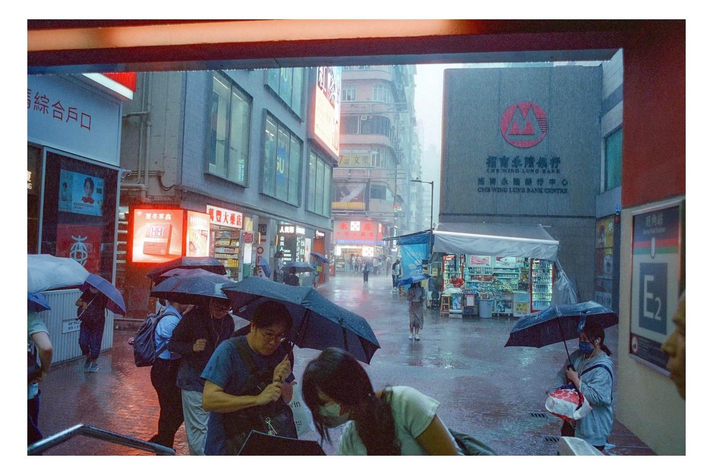 People with umbrellas walk along a wet street in a city during a rain shower.