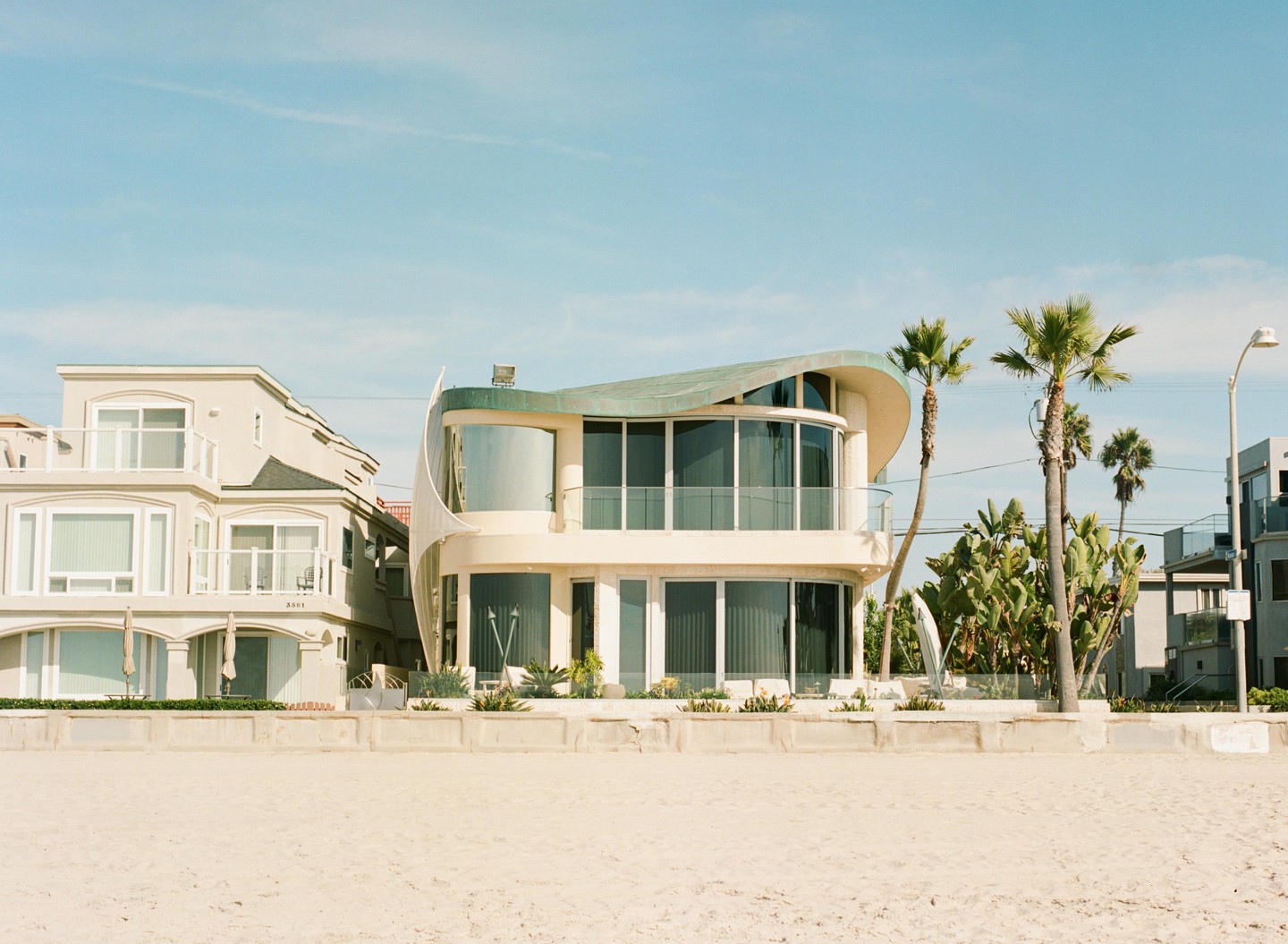 A modern beach house with a curved roof stands next to a traditional house on a sandy beach.