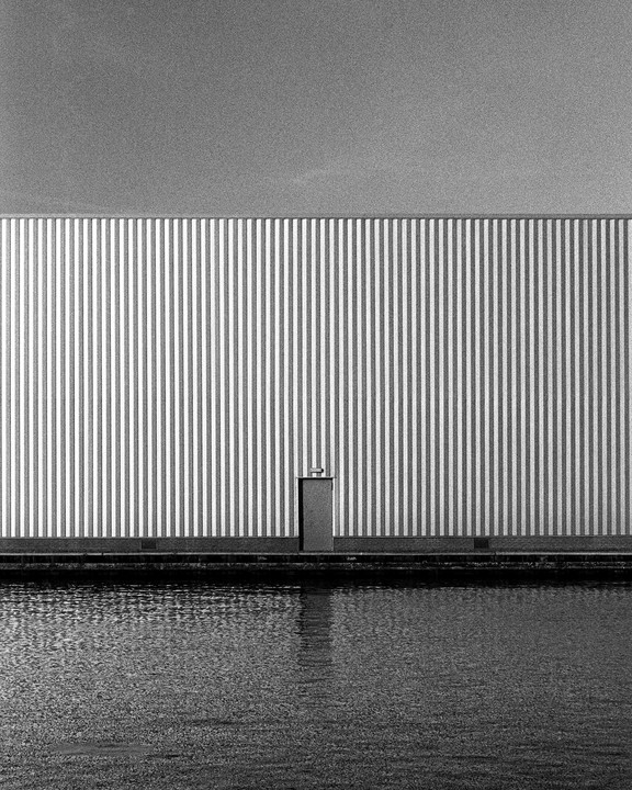 A minimalist composition features a striped wall with a small door next to a body of water under a clear sky.