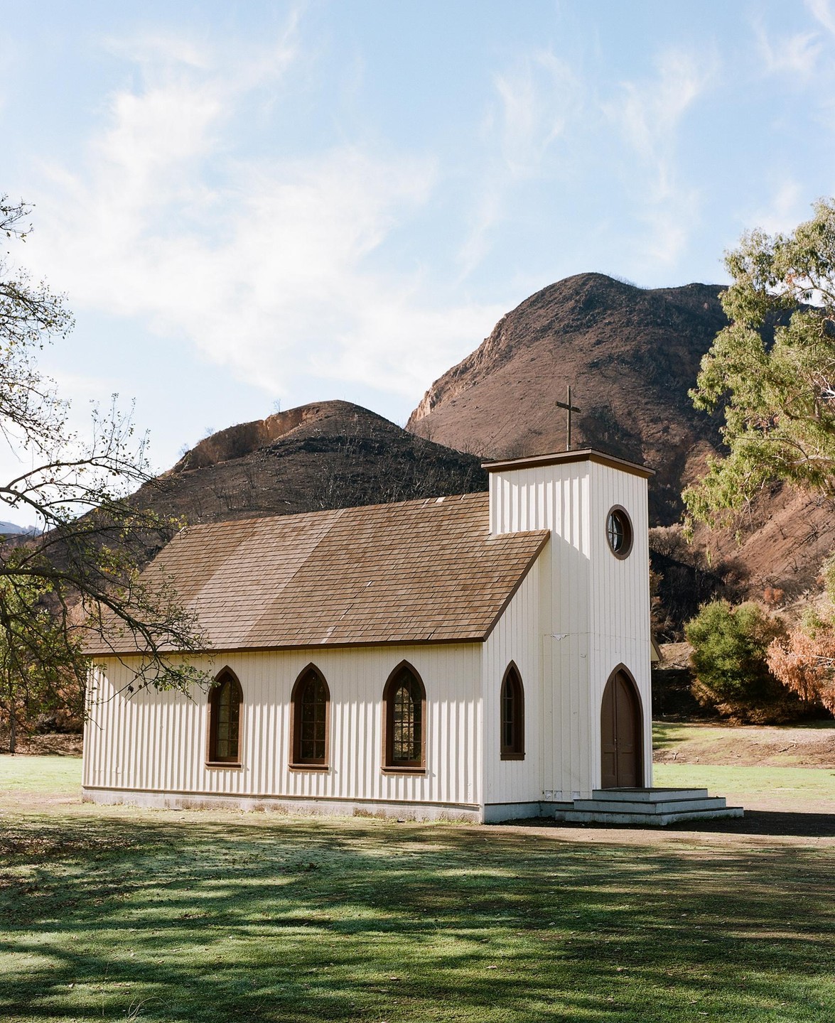 A white church with a brown roof stands in a grassy field with mountains in the background.