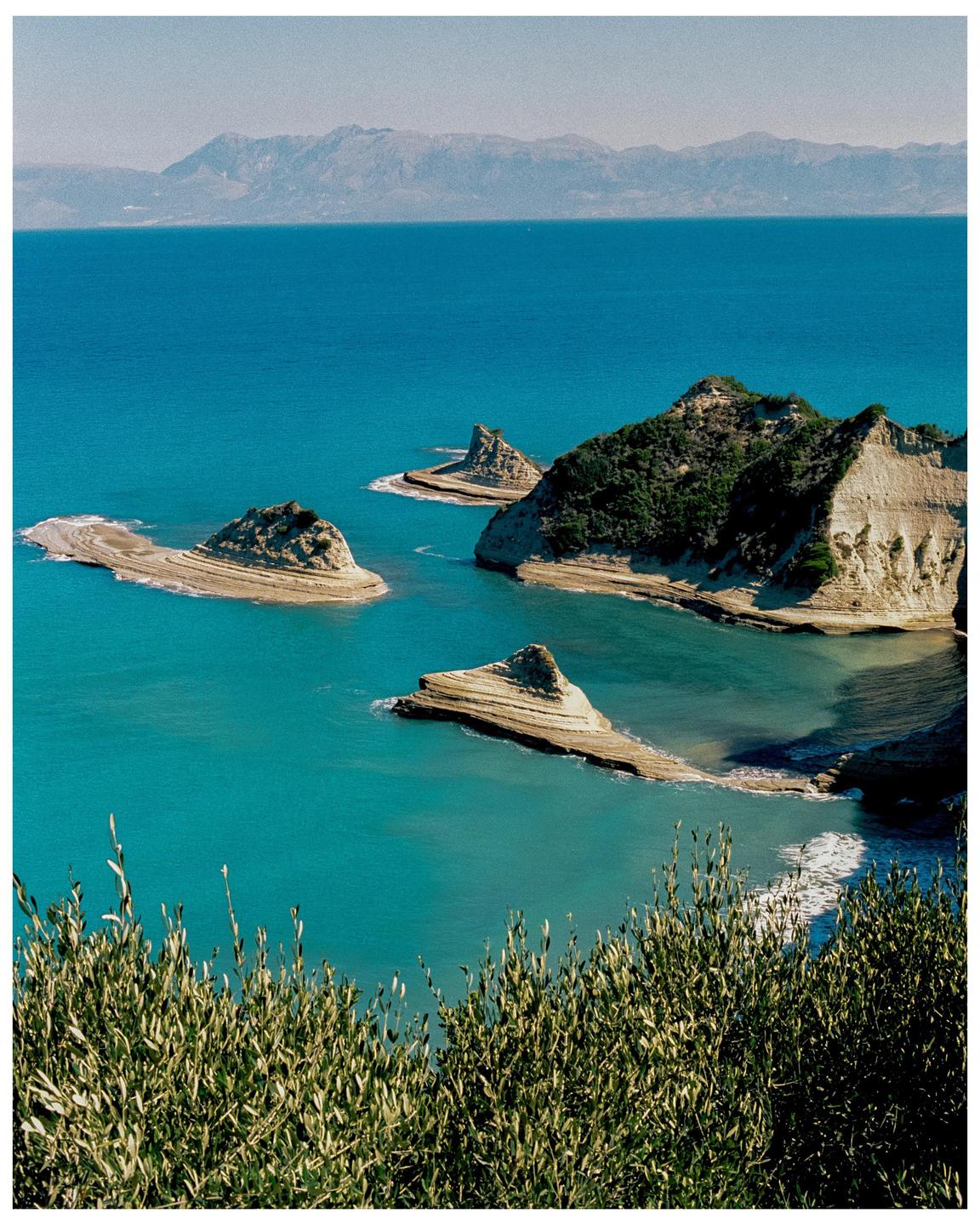 A high-angle landscape shows a bright blue sea with several small, rocky islands and a distant mountain range under a clear sky.