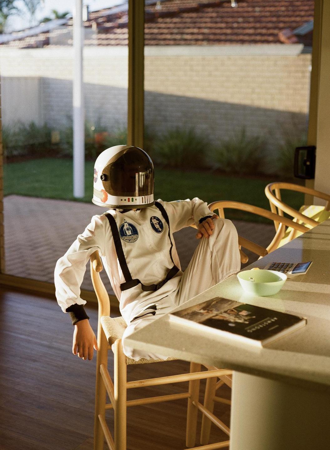 A child in an astronaut costume and helmet sits at a table with a book and a bowl, looking out a glass door to a yard.