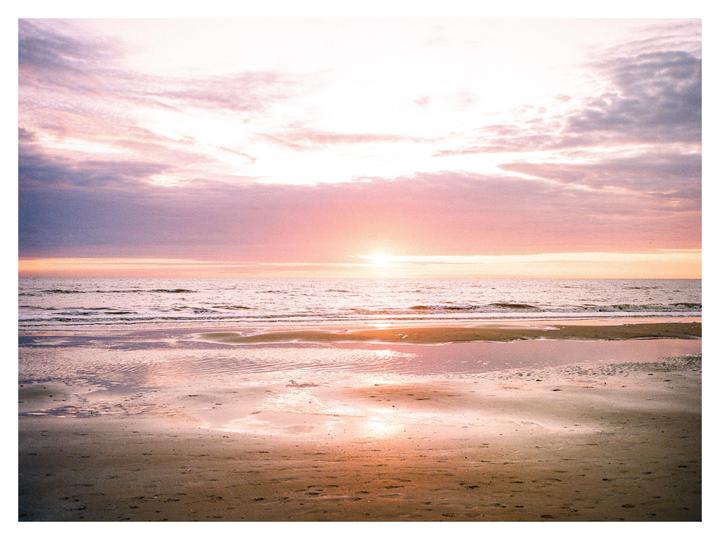 The sun sets over a beach with soft waves and colorful clouds.